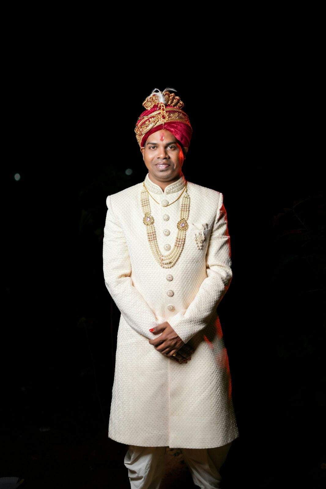 Man in traditional white sherwani with red turban against a black background