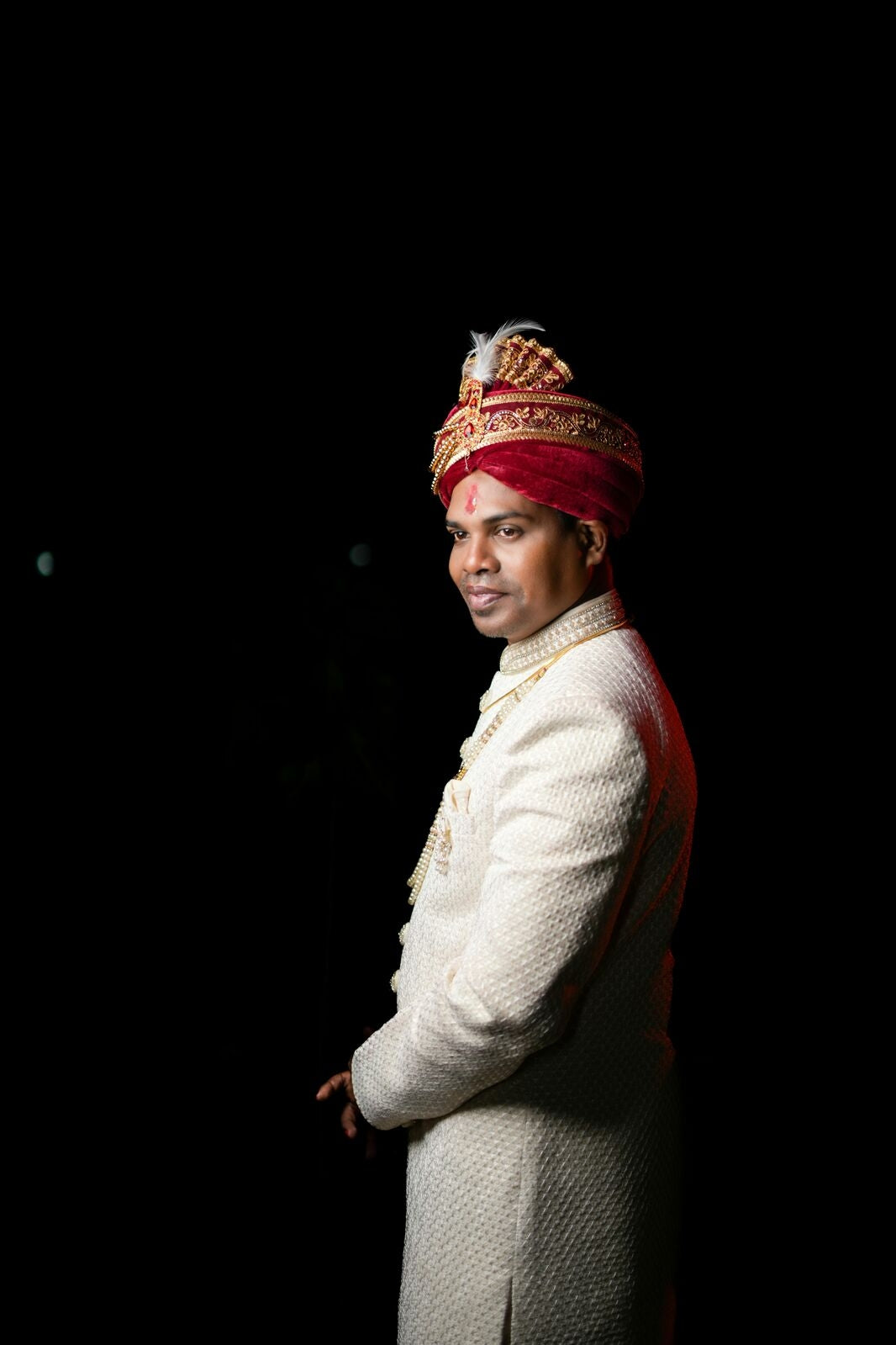 Man wearing a traditional white sherwani with a red turban against a black background